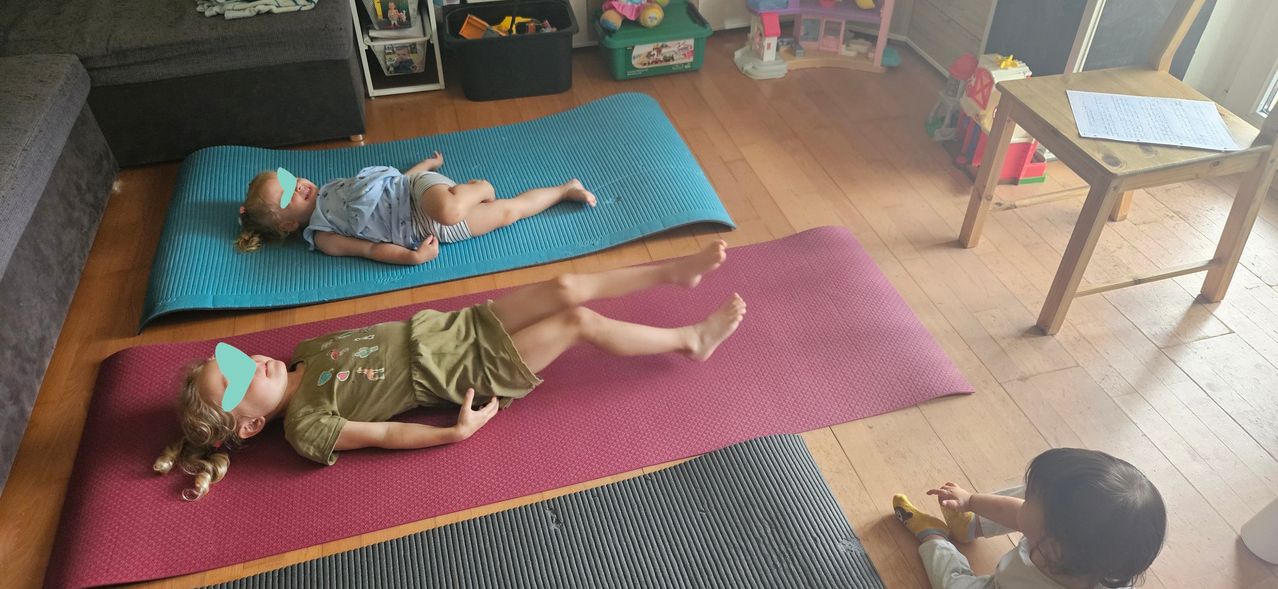 Two kids lie on blue and pink mats in a room with wooden floors, toys, and storage boxes. One kid is on a blue mat, the other on a pink mat.