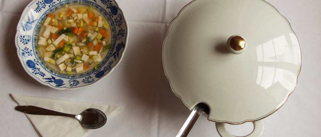 Eine Schüssel mit Suppe mit Gemüse- und Fleischstücken steht neben einem abgedeckten weißen Keramiktopf mit einem Löffel auf einer weißen Tischdecke.