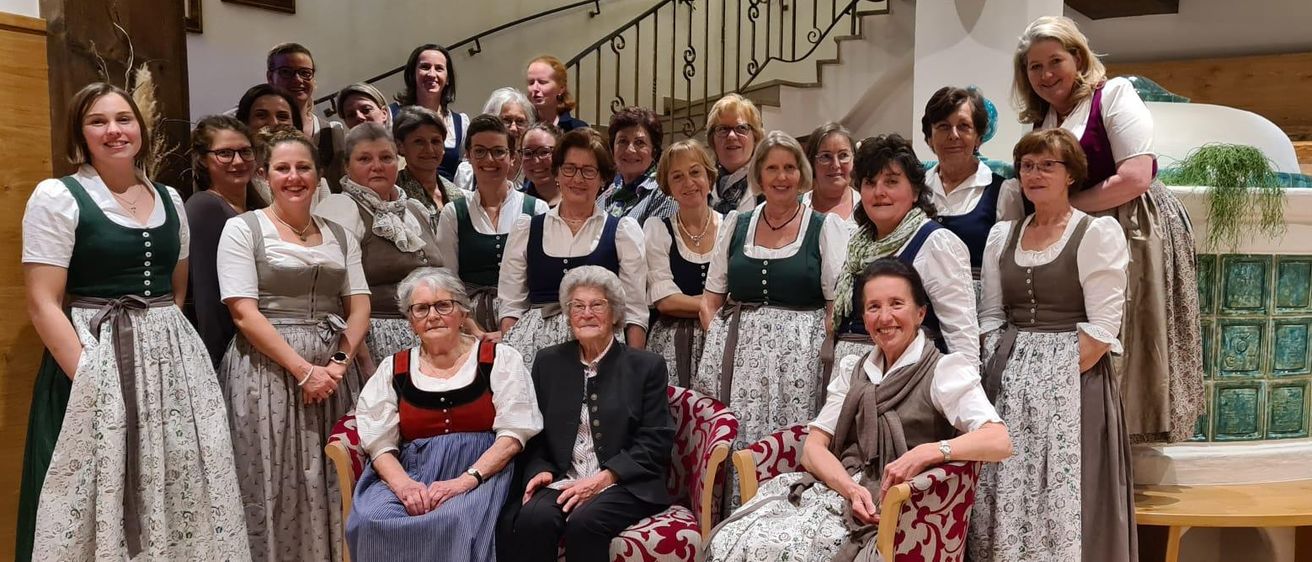 Eine Gruppe von Frauen in traditioneller Kleidung steht und sitzt auf Stühlen und lächelt. Dahinter ist eine Treppe zu sehen, und einige Frauen stehen in der Nähe.