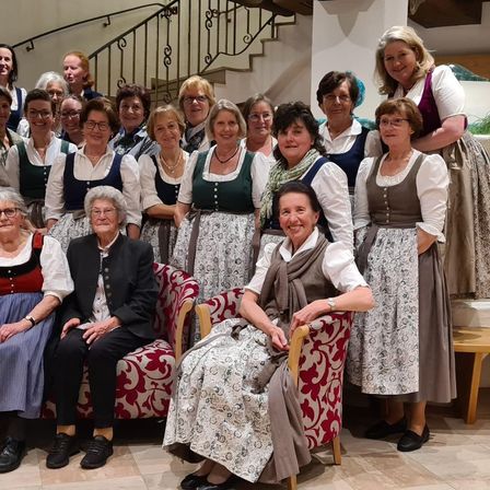 Eine Gruppe von Frauen, einige sitzend und andere stehend, in traditioneller Kleidung, posiert für ein Foto in einem Raum mit einer Treppe und einer Wand.