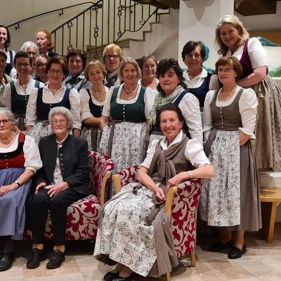 Eine Gruppe von Frauen, einige sitzend und andere stehend, in traditioneller Kleidung, posiert für ein Foto in einem Raum mit einer Treppe und einer Wand.