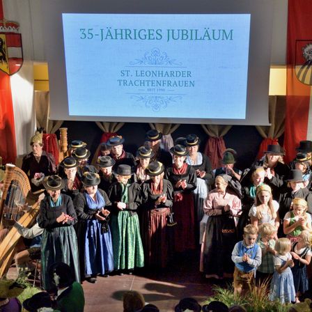 Ein Chor in traditionellen Kostümen steht auf der Bühne. Eine Projektionsleinwand hinter ihnen zeigt '35-jähriges Jubiläum'. Das Publikum sitzt vor ihnen.
