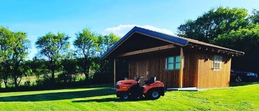 Ein kleines Holzhaus ist von saftigem grünem Gras umgeben. Vor ihm steht ein roter Rasenmäher. Der Himmel ist blau mit weißen Wolken.