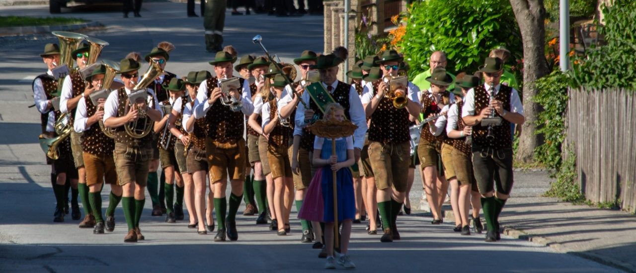 Eine Blaskapelle in traditioneller bayerischer Kleidung, einschließlich grüner Hüte und Westen, geht mit einem jungen Mädchen an der Spitze die Straße entlang.