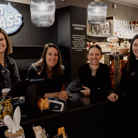 Vier Frauen sitzen an einem Tresen in einem Geschäft. Sie lächeln und schauen in die Kamera. Dahinter befindet sich eine Tafel mit einem Logo und einem Schild.