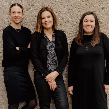 Drei Frauen in schwarzer Kleidung stehen gemeinsam vor einer Betonwand, lächeln und posieren für ein Foto.