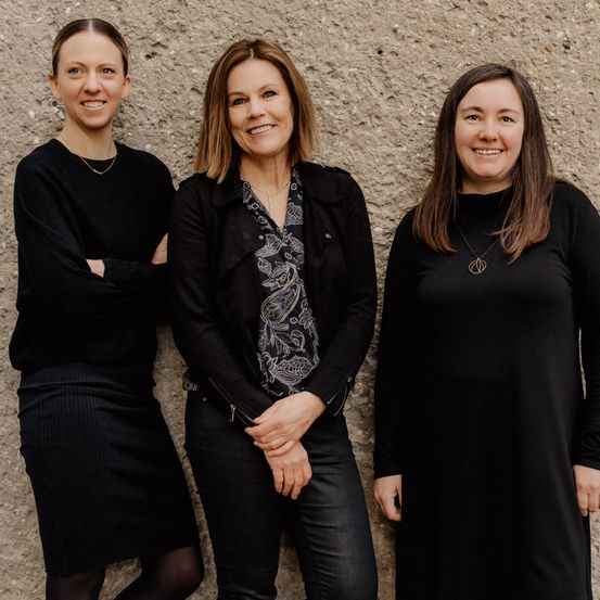 Drei Frauen in schwarzer Kleidung stehen gemeinsam vor einer Betonwand, lächeln und posieren für ein Foto.