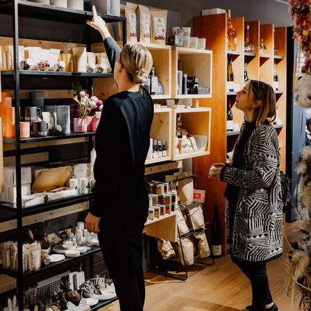 Zwei Frauen in einem Boutique, eine zeigt auf Artikel in einem Regal, während die andere aufschaut und einen Becher trägt.