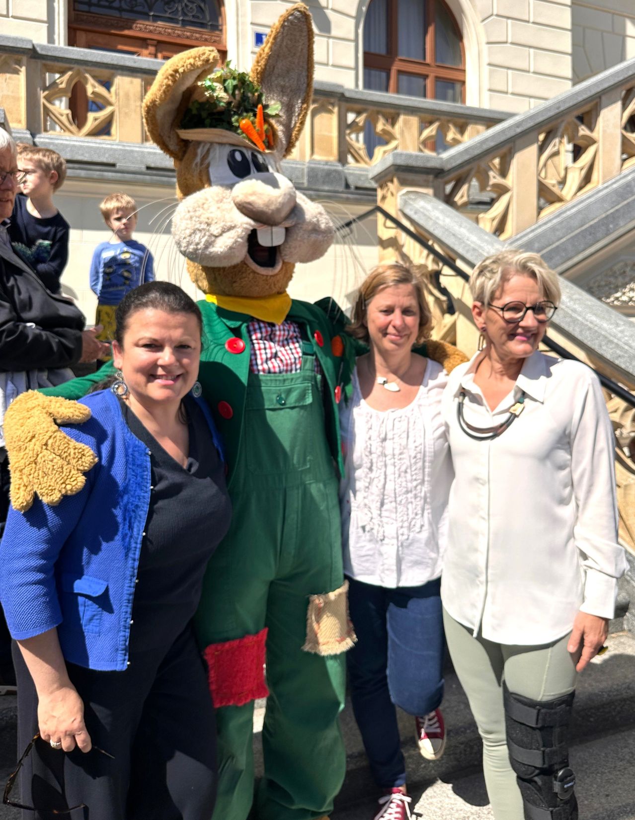 Drei Frauen und ein Mann posieren mit einem Maskottchen, das eine Karotte auf dem Kopf hat. Das Maskottchen trägt ein grünes Outfit und hat ein lächelndes Gesicht. Die Frauen lächeln und scheinen für ein Foto zu posieren. Der Mann hinter ihnen schaut sich etwas an.