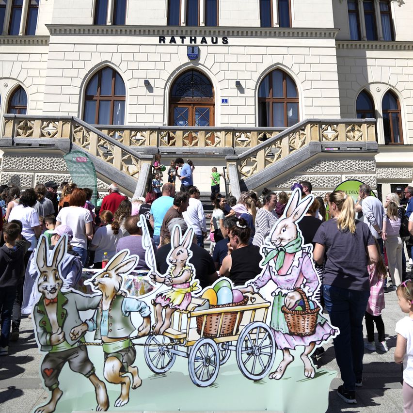 Menschen versammeln sich vor einem großen Gebäude mit einem gewölbten Eingang und einem Schild, das 'Rathaus' steht. Im Vordergrund befindet sich eine große Ausschnitt von Hasen, die einen Korb ziehen.