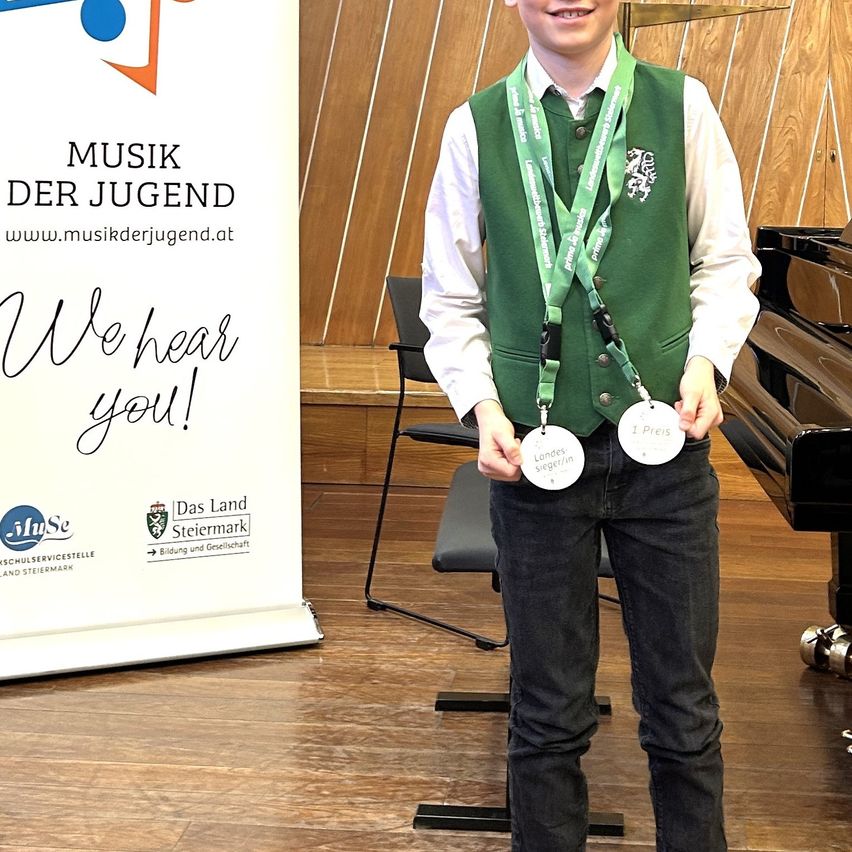 Ein Junge steht vor einer Holzwand mit einem Musikbanner. Er trägt eine grüne Weste und hält eine weiße Medaille mit der Aufschrift '1 Preis'.