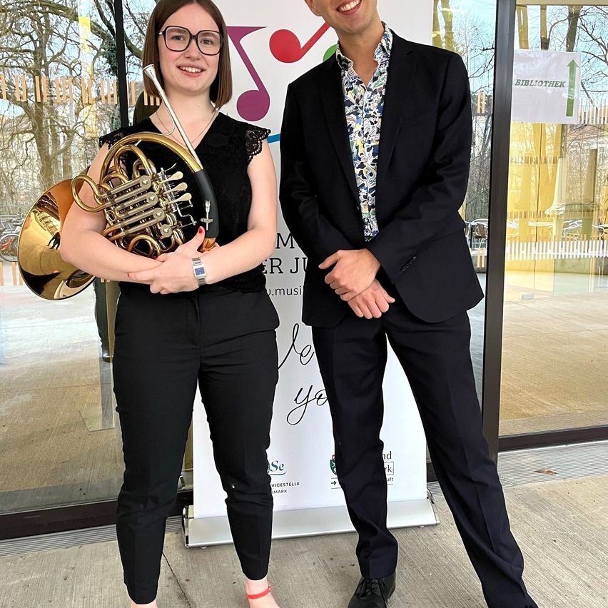 Zwei Personen stehen vor einem Banner mit Noten und Text. Eine Frau hält ein Horn und lächelt. Ein Mann neben ihr trägt einen Anzug und schaut in die Kamera.