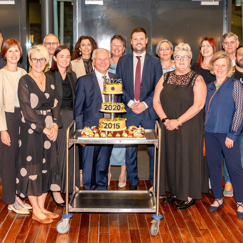 Eine Gruppe von Menschen posiert für ein Foto, wobei zwei Männer vor einem Wagen stehen, der einen Kuchen mit den Jahren 2025, 2020 und 2015 darauf hält.