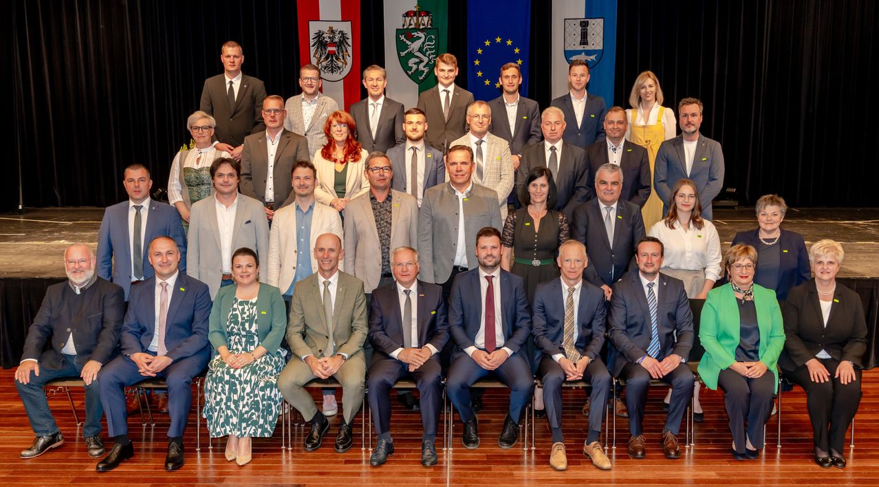 Eine Gruppe Erwachsener in formeller Kleidung posiert vor drei Flaggen zu einem Foto.