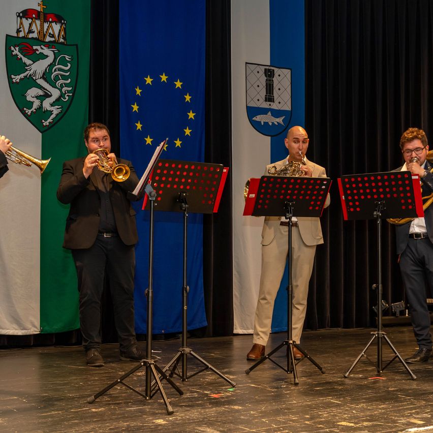 Vier Männer spielen Trompeten und ein Waldhorn auf einer Bühne mit einem Hintergrund von Flaggen aus Italien, der Europäischen Union und einer Stadt.