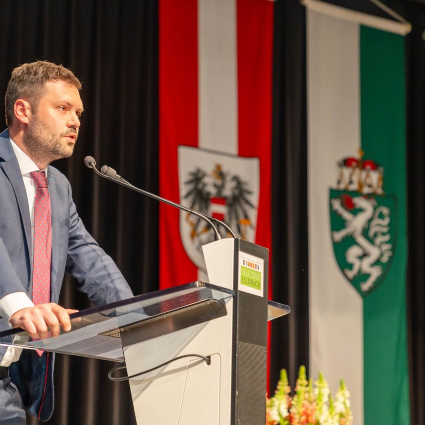 Ein Mann in einem Anzug steht an einem Podium mit Mikrofonen und hält eine Rede. Hinter ihm befinden sich zwei Flaggen, eine mit einem Löwenwappen und die andere mit einem Schildwappen.