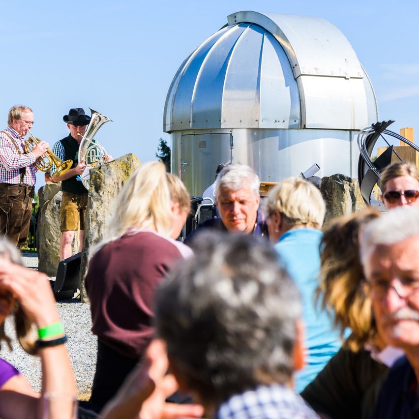 Ein Mann spielt neben einem Teleskop eine Trompete. Dahinter spielen zwei Männer in traditioneller deutscher Kleidung eine Tuba. Ein Publikum älterer Erwachsener ist vor ihnen versammelt.