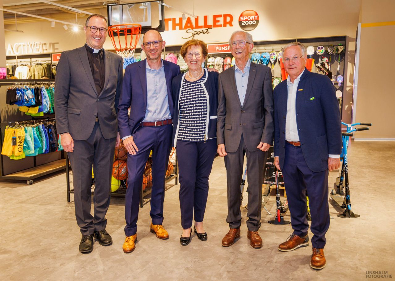 Fünf Menschen in formeller Kleidung posieren für ein Foto vor einem Sportgeschäft mit dem Schild Thaller Sport 2000. Die Frau trägt eine Halskette und eine Brille.