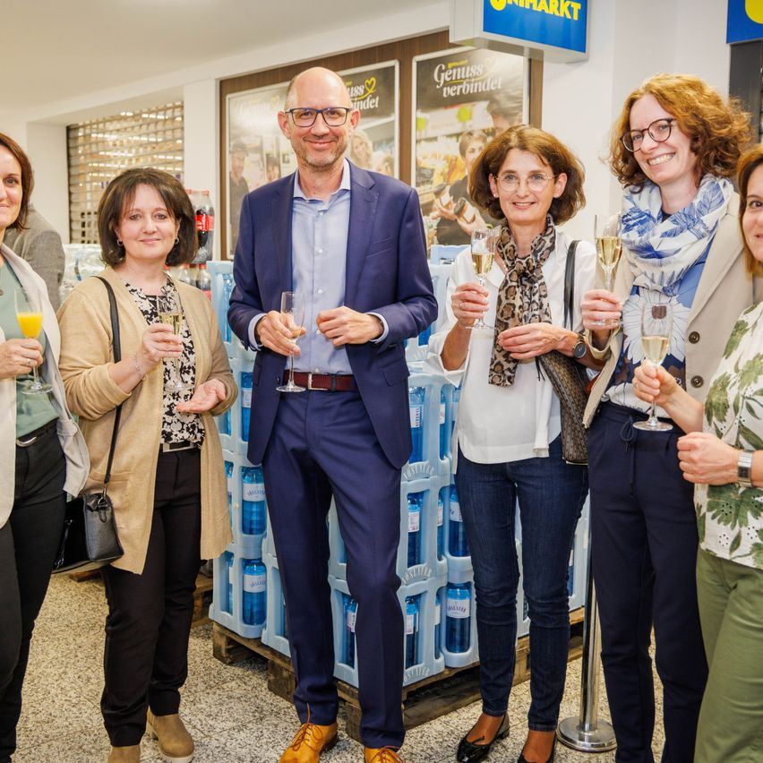 Eine Gruppe von Erwachsenen posiert für ein Foto in einem Geschäft mit Produkten im Hintergrund. Sie alle lächeln und halten Weingläser mit Getränken.