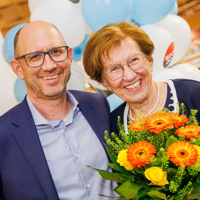 Ein Mann und eine ältere Frau lächeln vor einer Wand mit blauen und weißen Luftballons, möglicherweise zur Feier eines besonderen Anlasses.