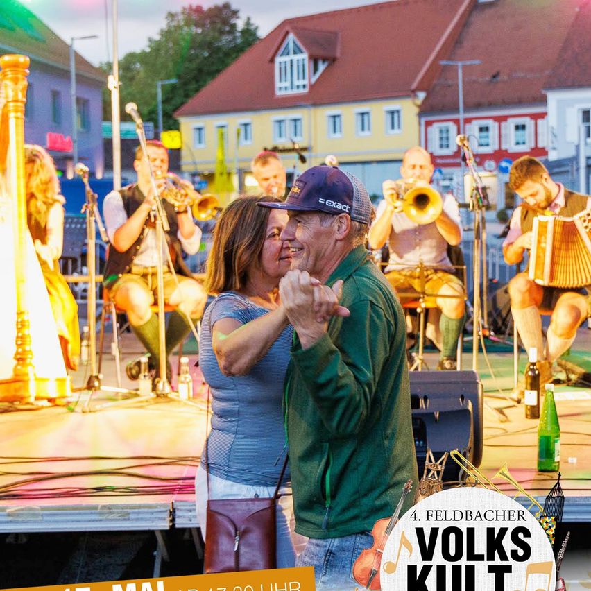 Ein Paar tanzt auf einer Bühne, während eine Band hinter ihnen spielt. Die Band besteht aus Trompete, Akkordeon und Harfe. Die Bühne ist gut beleuchtet, und im Hintergrund sind Häuser und Bäume zu sehen.