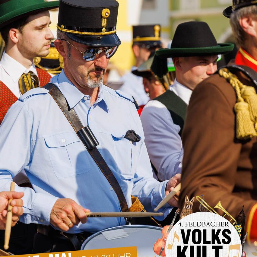 Eine Gruppe von Männern in Uniform, darunter einer, der eine Trommel spielt, nehmen an einer Parade teil. Sie tragen Hüte und einige haben Sonnenbrillen. Das Bild enthält Text auf Deutsch.