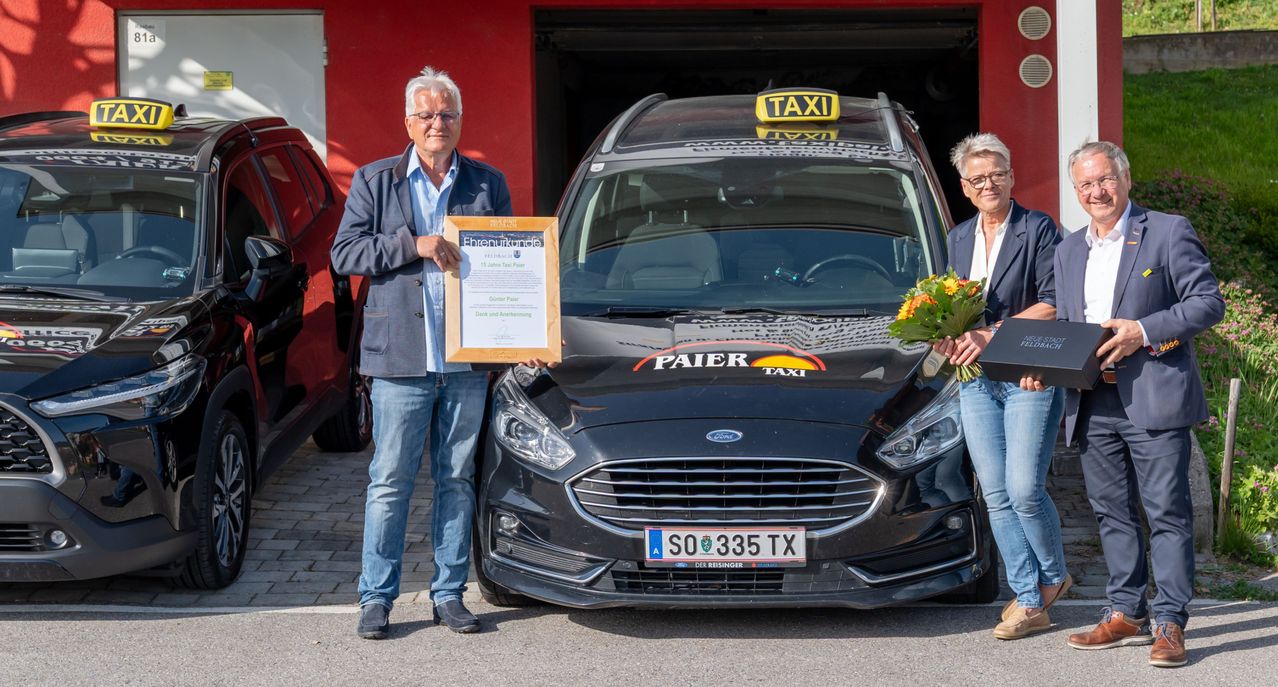 Ein Mann mit einem Zertifikat steht neben einem Taxi, beide lächeln. Das Taxi hat ein gelbes Schild oben und einen Blumenstrauß an der Seite.