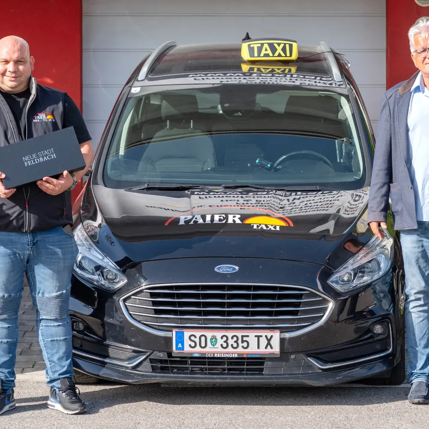 Zwei Männer stehen neben einem schwarzen Ford-Taxi, einer hält eine schwarze Box mit dem Text 'Neue Stadt Feldbach'. Das Taxi hat ein Taxischild oben und ein Nummernschild SO 335 TX.