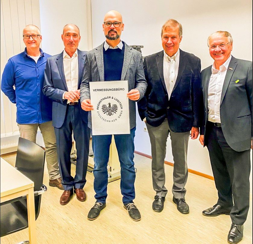 Fünf Männer stehen in einem Raum, einer hält ein Zertifikat. Sie sind alle in Anzügen gekleidet. Ein Mann hat ein weißes Schild mit deutschem Text.