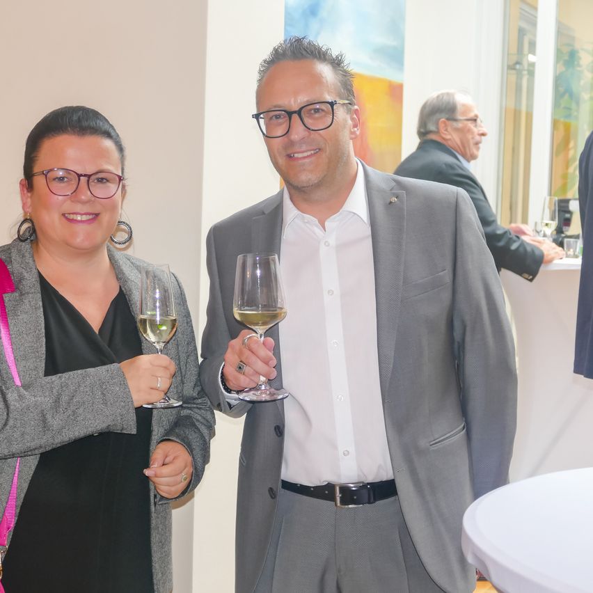 Zwei Personen in formeller Kleidung lächeln und halten Weinpokale, vielleicht bei einer gesellschaftlichen Zusammenkunft. Hinter ihnen sitzt ein Mann an einem Tisch mit einem Becher. Eine weiße Wand und ein Glasfenster sind im Hintergrund.