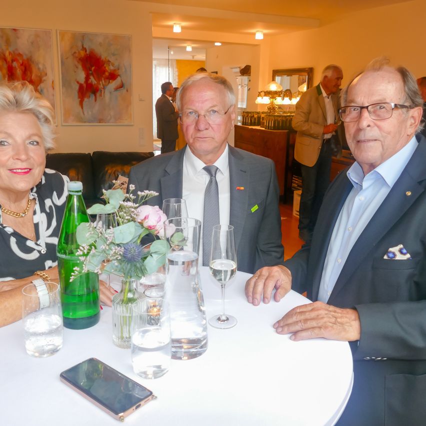Drei Menschen in Anzügen sitzen um einen Tisch mit Gläsern, Blumen und einem Handy. Sie sind in einem Raum mit einer Couch, Gemälden und einem anderen Mann.