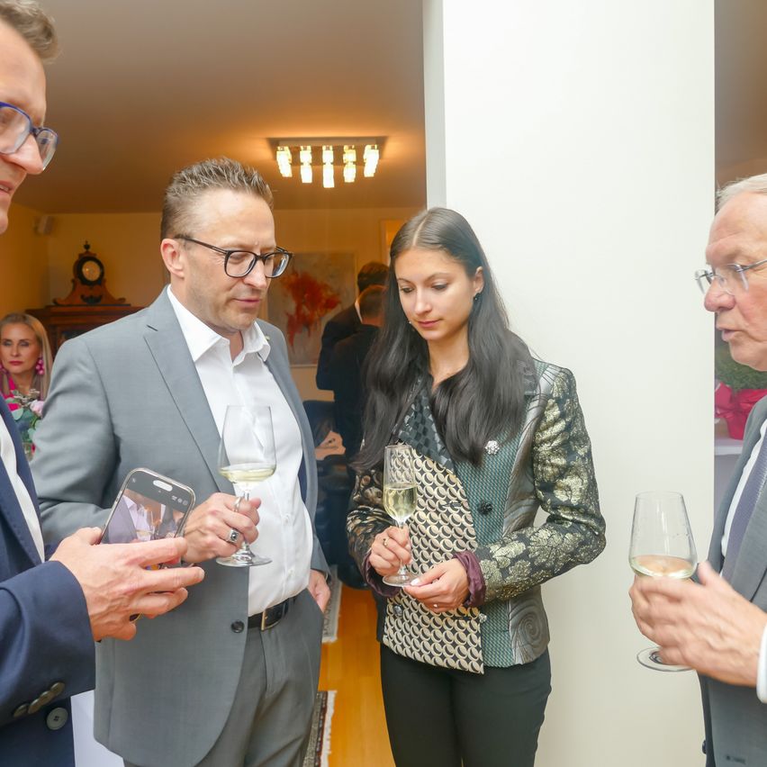 Drei Menschen in formeller Kleidung stehen in einem Raum und halten Weingläser. Ein Mann links schaut auf sein Handy. Eine Frau in der Mitte hält ein Glas Wein. Ein älterer Mann rechts schaut etwas an.