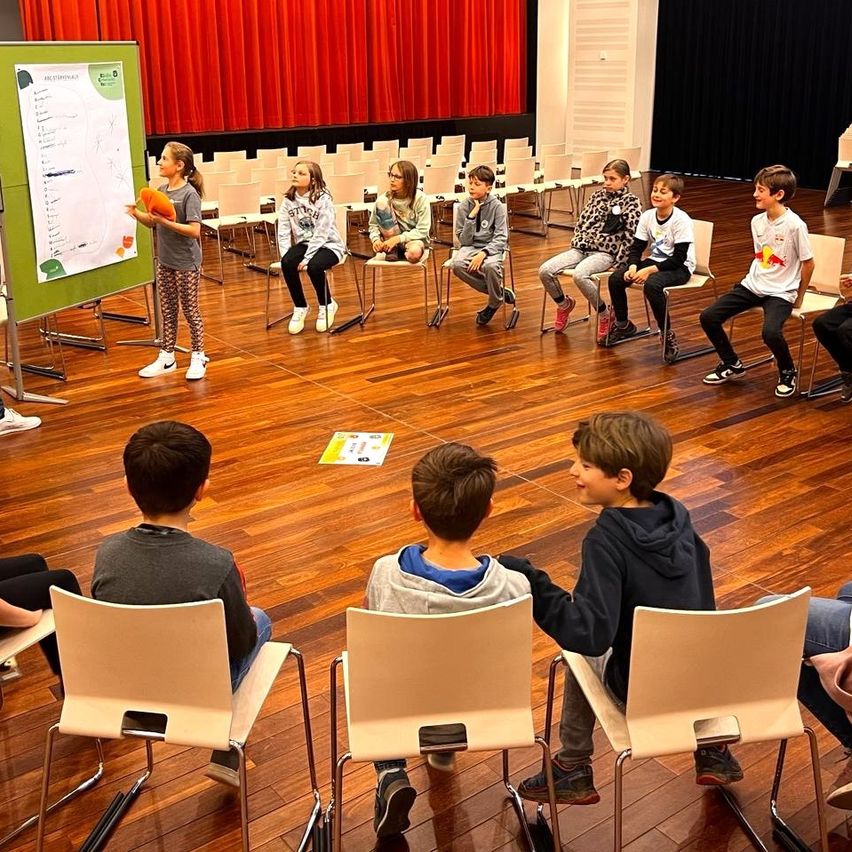 Eine Gruppe von Kindern sitzt in einem Kreis in einem Raum und hört einem Mädchen zu, das vor ihnen steht und ein Objekt in der Hand hält.