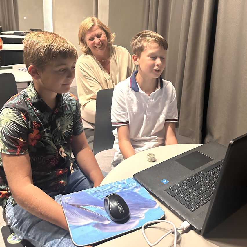 Zwei Jungen sitzen an einem Tisch mit einem Laptop, einer trägt ein Blumenhemd, der andere ein weißes Hemd. Eine lächelnde Frau sitzt hinter ihnen.
