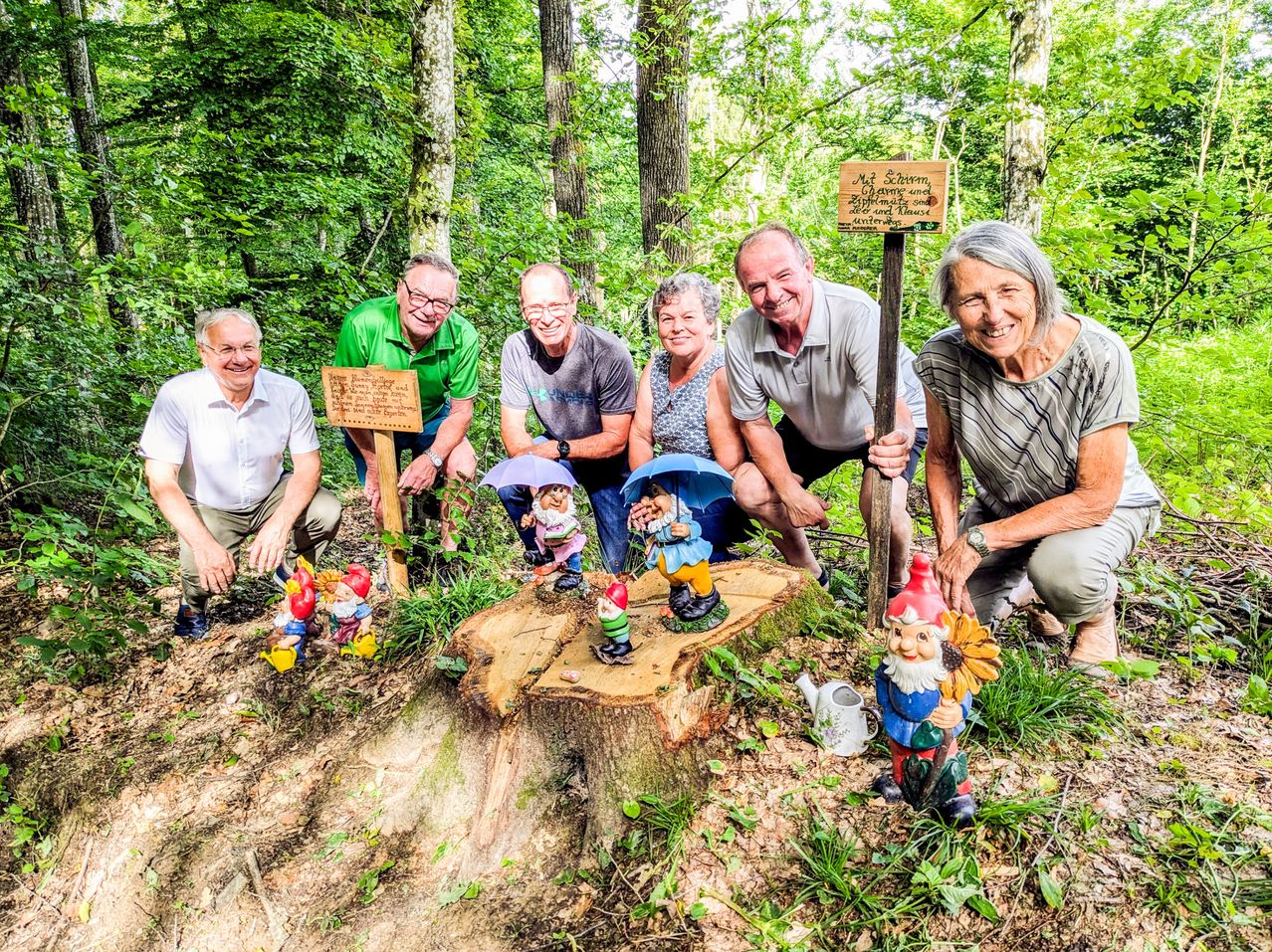 Sechs Personen posieren in einem Wald mit Gnom-Statuen, einem Holzschild und einer Gießkanne. Sie lächeln und sehen glücklich aus. Der Wald ist voller Bäume und Pflanzen.