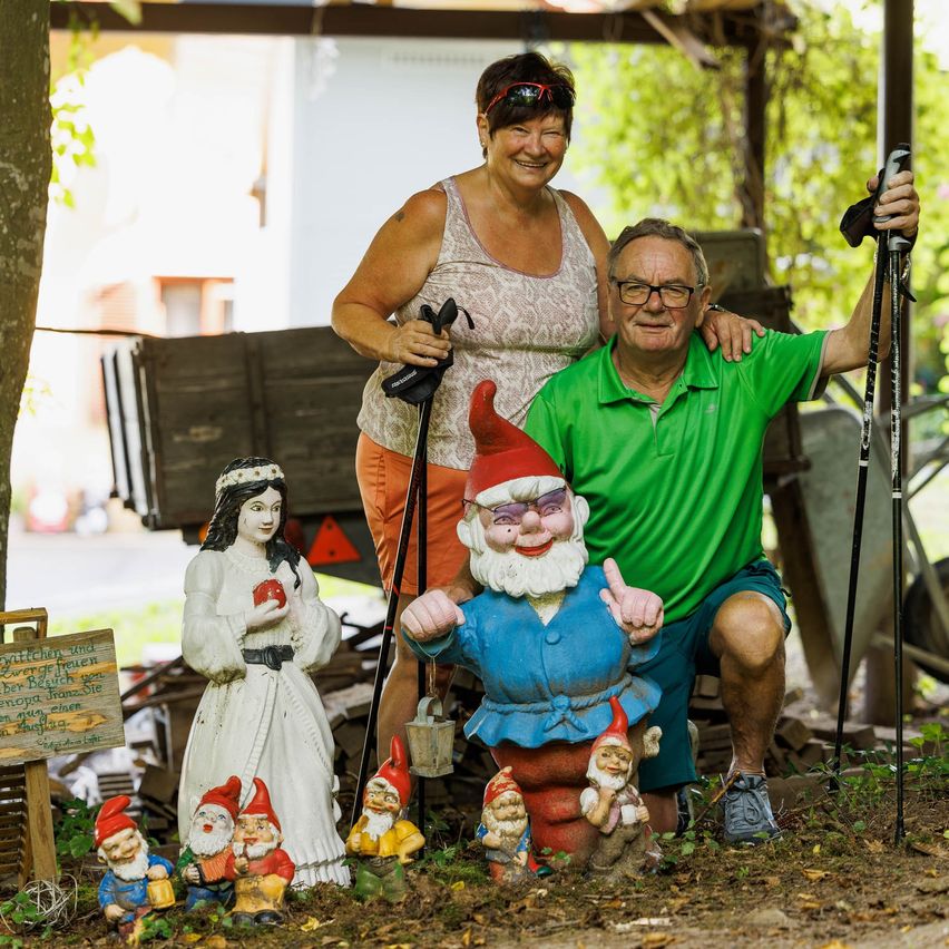 Ein älteres Paar posiert für ein Foto mit Gartenzwerg-Figuren. Die Frau lächelt und trägt eine Sonnenbrille auf dem Kopf. Der Mann hält einen Gehstock und ein Schild. Die Zwerge stehen auf dem Boden und halten einige Objekte. Im Hintergrund befinden sich Holzkonstruktionen und Bäume.