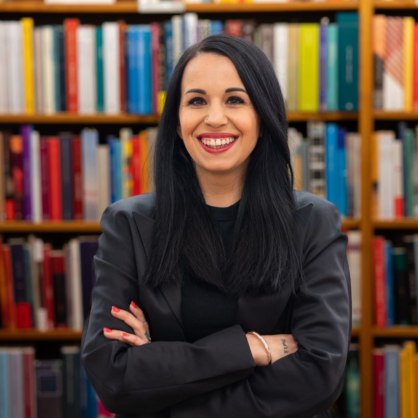 Eine lächelnde Frau mit langen schwarzen Haaren steht vor einem Bücherregal, das mit Büchern in verschiedenen Farben gefüllt ist. Sie trägt einen schwarzen Blazer und hat die Arme verschränkt.