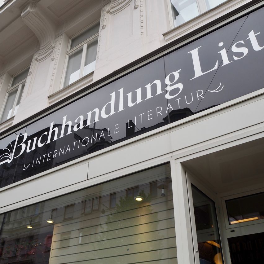Ein Ladenschild für ein Buchgeschäft namens 'Buchhandlung List'. Es ist in Weiß auf dunklem Hintergrund geschrieben, mit 'INTERNATIONALE LITERATUR' darunter. Der Laden ist modern, mit einer Glastür und reflektierenden Fenstern.