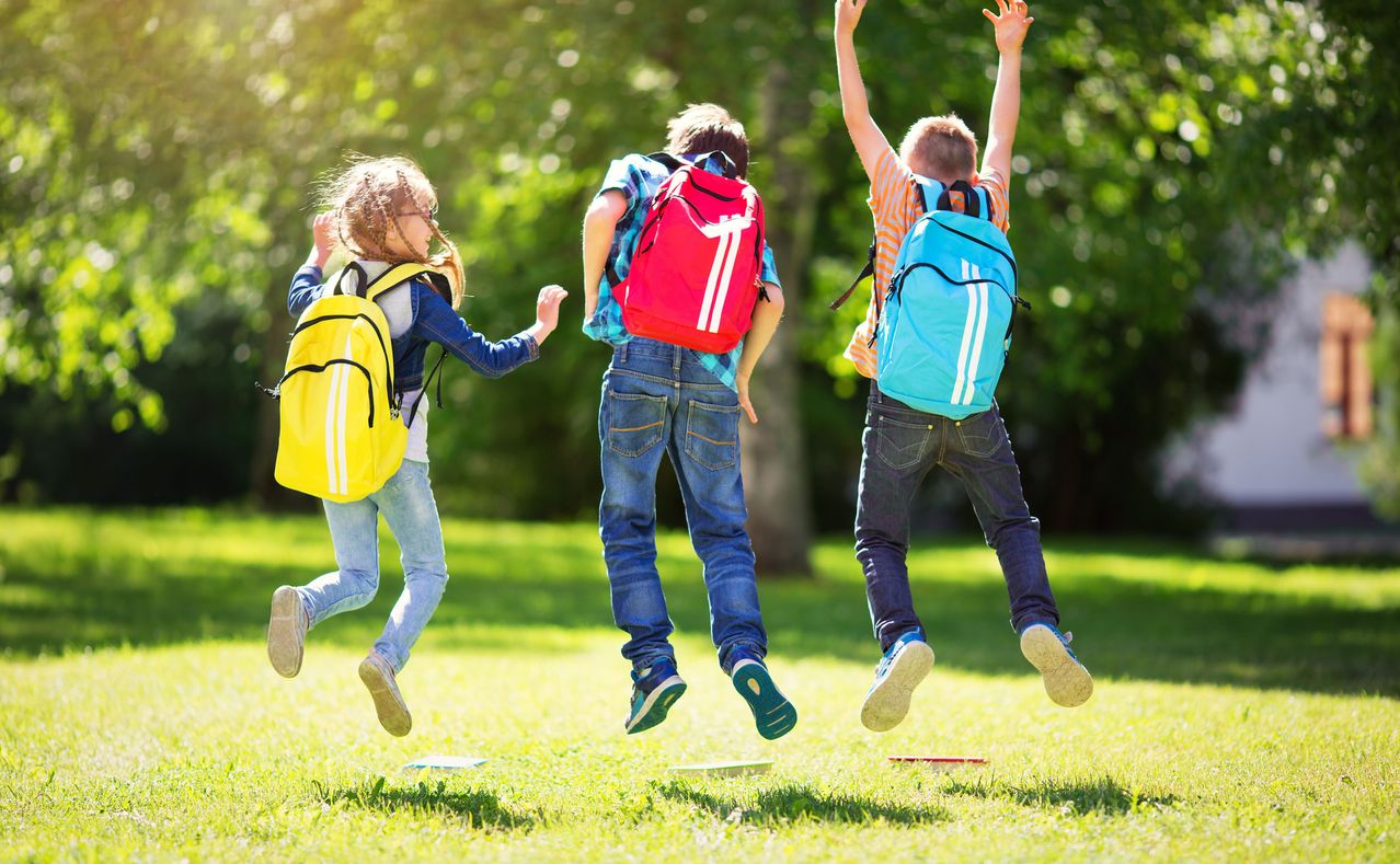 Drei Kinder springen an einem sonnigen Tag in einem Park in die Luft. Jedes Kind hat einen Rucksack auf dem Rücken. Das Mädchen trägt einen gelben Rucksack, der Junge in der Mitte einen roten und der Junge auf der rechten Seite einen blauen.