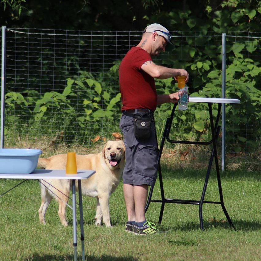 Ein Mann gießt ein Getränk in einen Becher auf einem Tisch, während ein gelber Labrador neben ihm steht. Sie befinden sich in einem eingezäunten Außenbereich mit Grünpflanzen im Hintergrund.