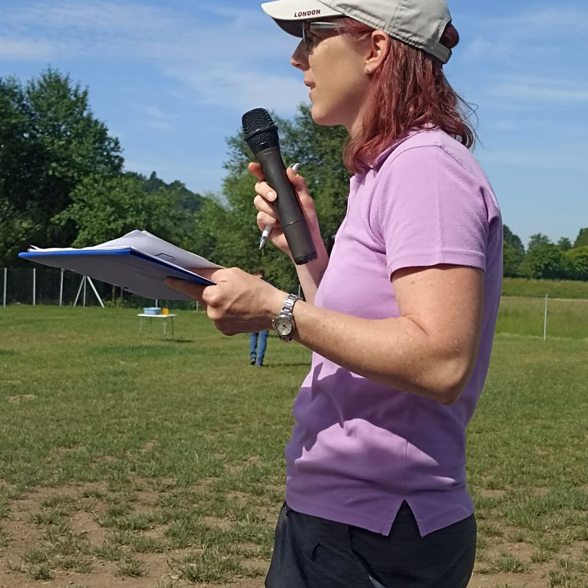 Eine Frau in einem lila Shirt hält ein Mikrofon und schaut nach vorne, steht in einem Grasfeld mit einem Tisch im Hintergrund.