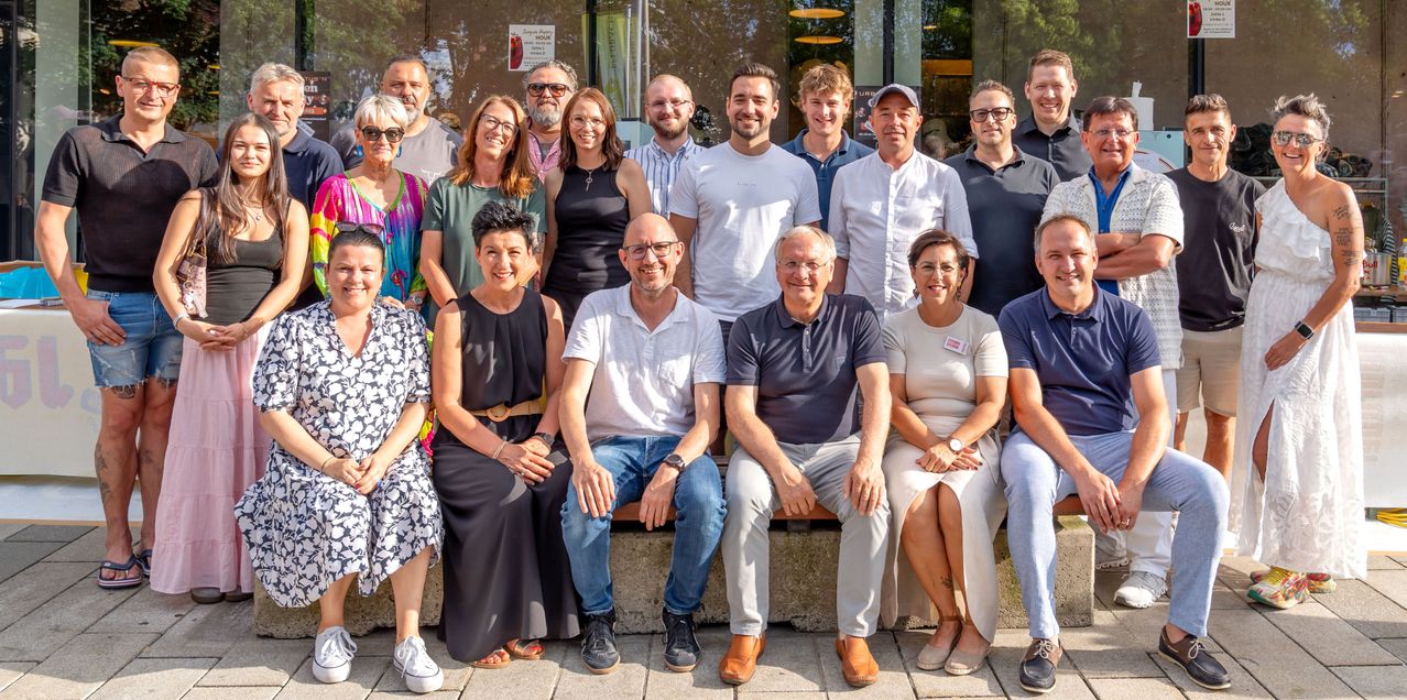 Eine Gruppe von Menschen, sowohl Männer als auch Frauen, versammelt sich für ein Foto. Sie stehen und sitzen auf einer Betonbank vor einem Gebäude. Sie alle lächeln und tragen Freizeitkleidung. Hinter ihnen befindet sich eine Glaswand mit einem Poster.