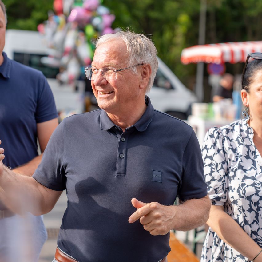 Ein älterer Mann mit Brille und blauem Poloshirt lächelt und gestikuliert neben einer Frau in einem Blumenkleid. Im Hintergrund befinden sich verschwommen ein weißer Van und ein rot-weiß gestreifter Sonnenschirm.