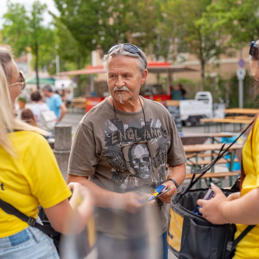 Ein älterer Mann mit Schnurrbart und Sonnenbrille auf dem Kopf unterhält sich mit zwei Frauen in einem Park.
