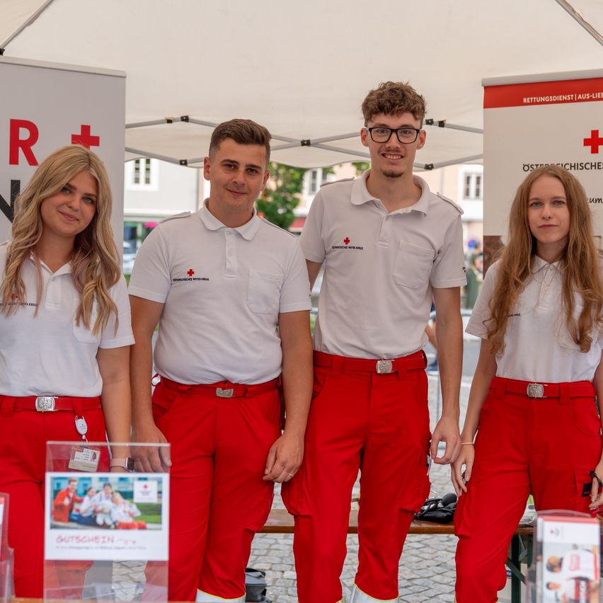Vier lächelnde Freiwillige in weißen und roten Uniformen stehen zusammen unter einem Zelt mit einem roten Kreuzbanner. Eine Frau links hat blonde Haare, während die anderen Männer sind.