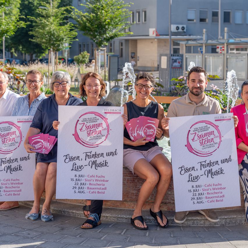 Eine Gruppe von Menschen posiert für ein Foto in einer Außenumgebung. Sie halten Schilder mit dem Text 'Essen, Trinken und Live-Musik'. Die Menschen tragen Freizeitkleidung und lächeln. Hinter ihnen steht ein Brunnen mit aufspritzendem Wasser. Im Hintergrund befindet sich ein Gebäude mit Fenstern und einem Baum.