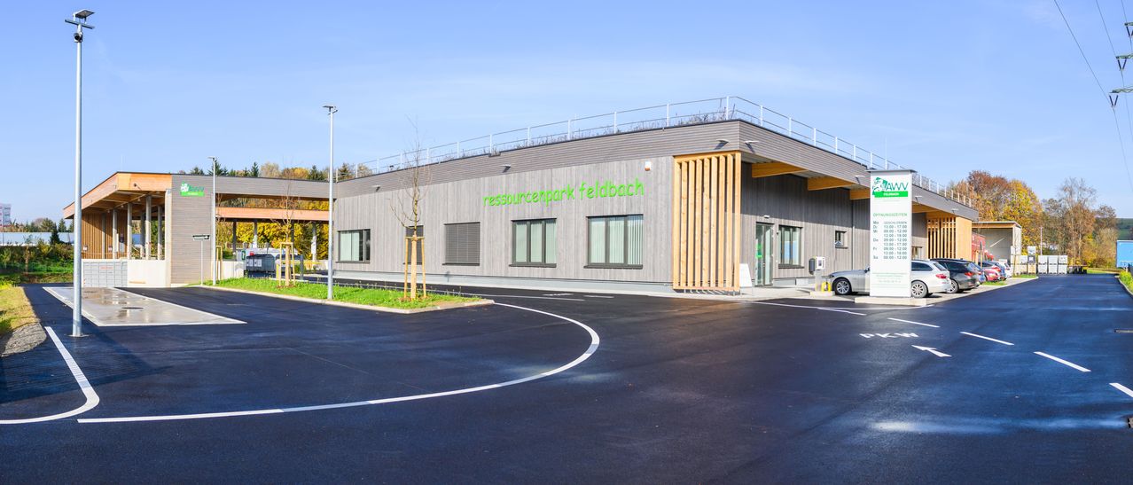 Das Bild zeigt ein modernes, graues Gebäude mit der Aufschrift 'ressourcenpark feldbach'. Das Gebäude verfügt über eine Dachterrasse, Glasfenster und hölzerne Eingangstüren. Vor dem Gebäude steht ein kleiner Baum, und davor befindet sich ein gepflasterter Parkplatz.