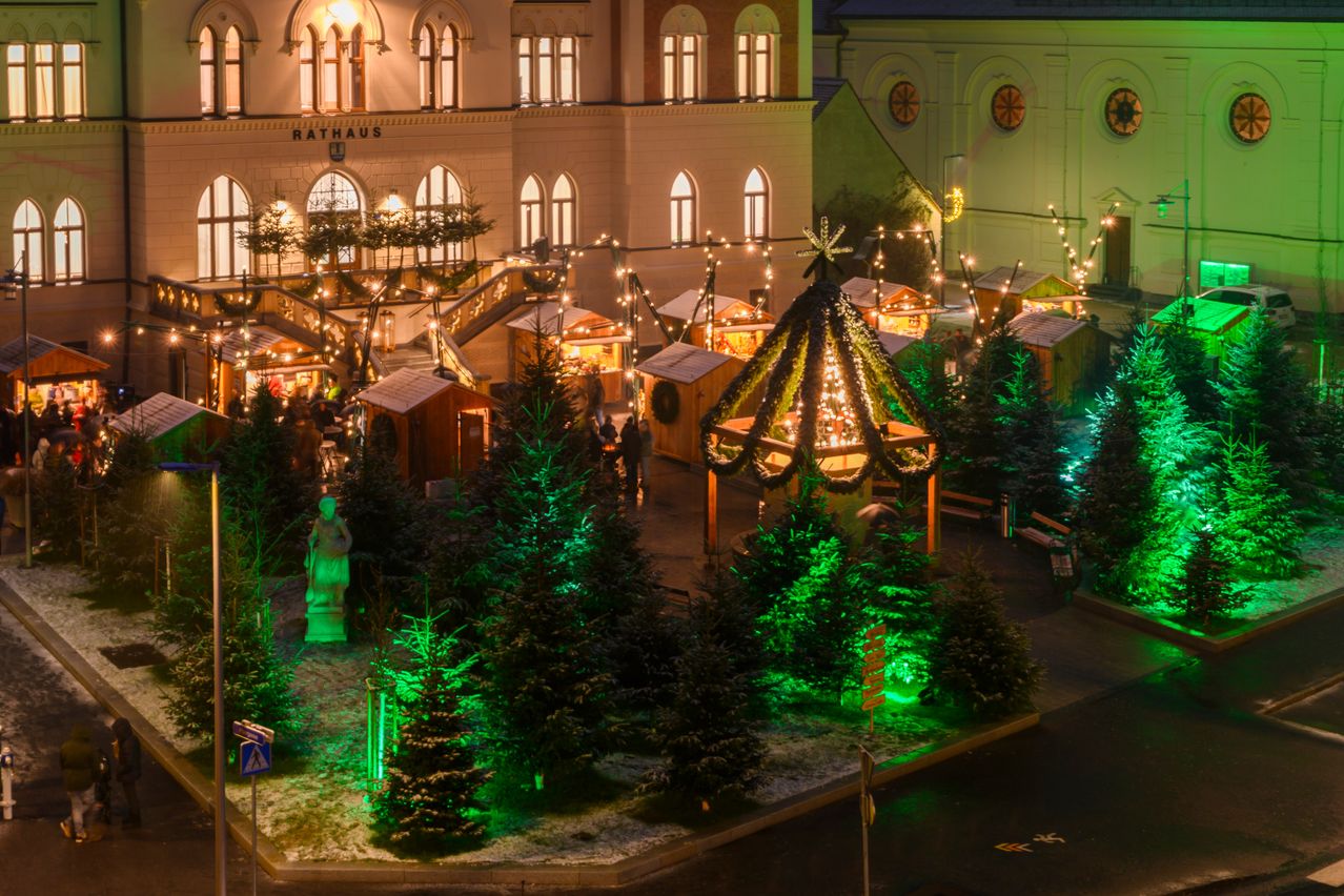 Ein Weihnachtsmarkt vor dem Rathausgebäude mit festlichen Lichtern, Holzbuden und grünen Bäumen, die mit grünen Lichtern beleuchtet sind.
