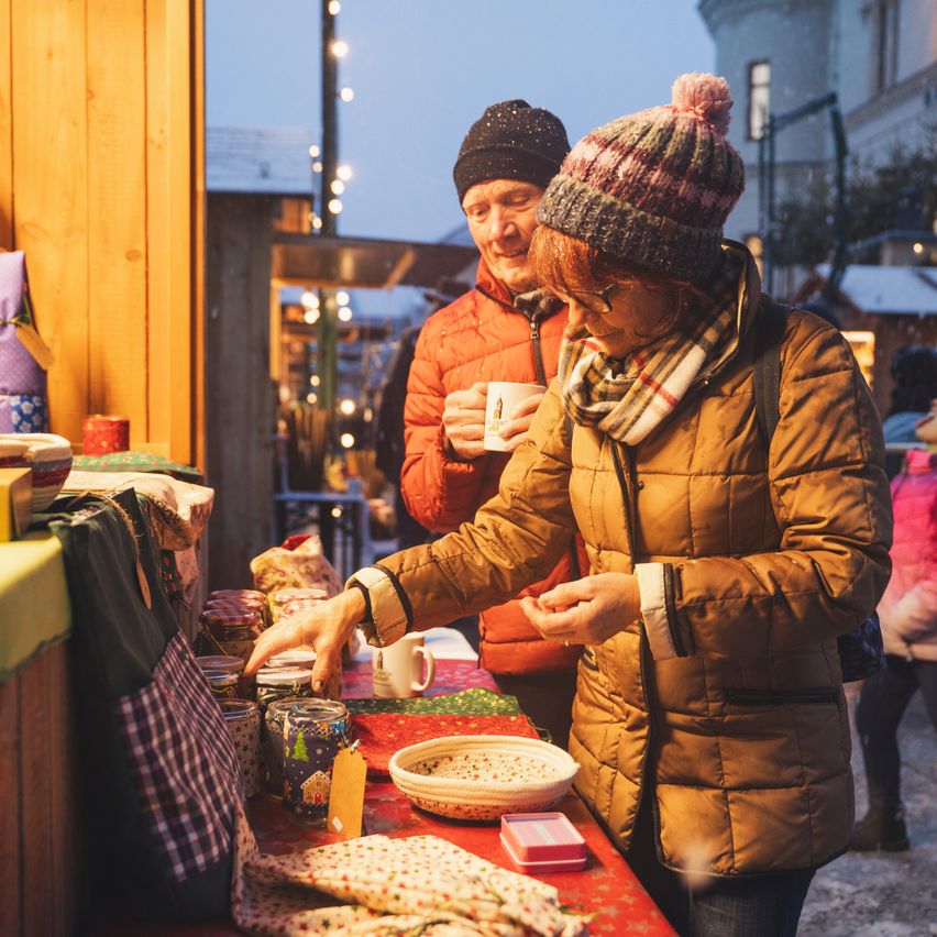 Zwei Personen kaufen auf einem Weihnachtsmarkt ein, wobei eine Frau Gegenstände auf einem Tisch betrachtet und eine Tasse in der Hand hält, während ein Mann hinter ihr steht.