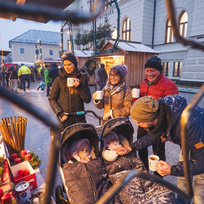 Eine Familie mit zwei Babys in Kinderwagen genießt Kaffee und Gespräche auf einem Weihnachtsmarkt, umgeben von festlichen Dekorationen und einer geschäftigen Menge.